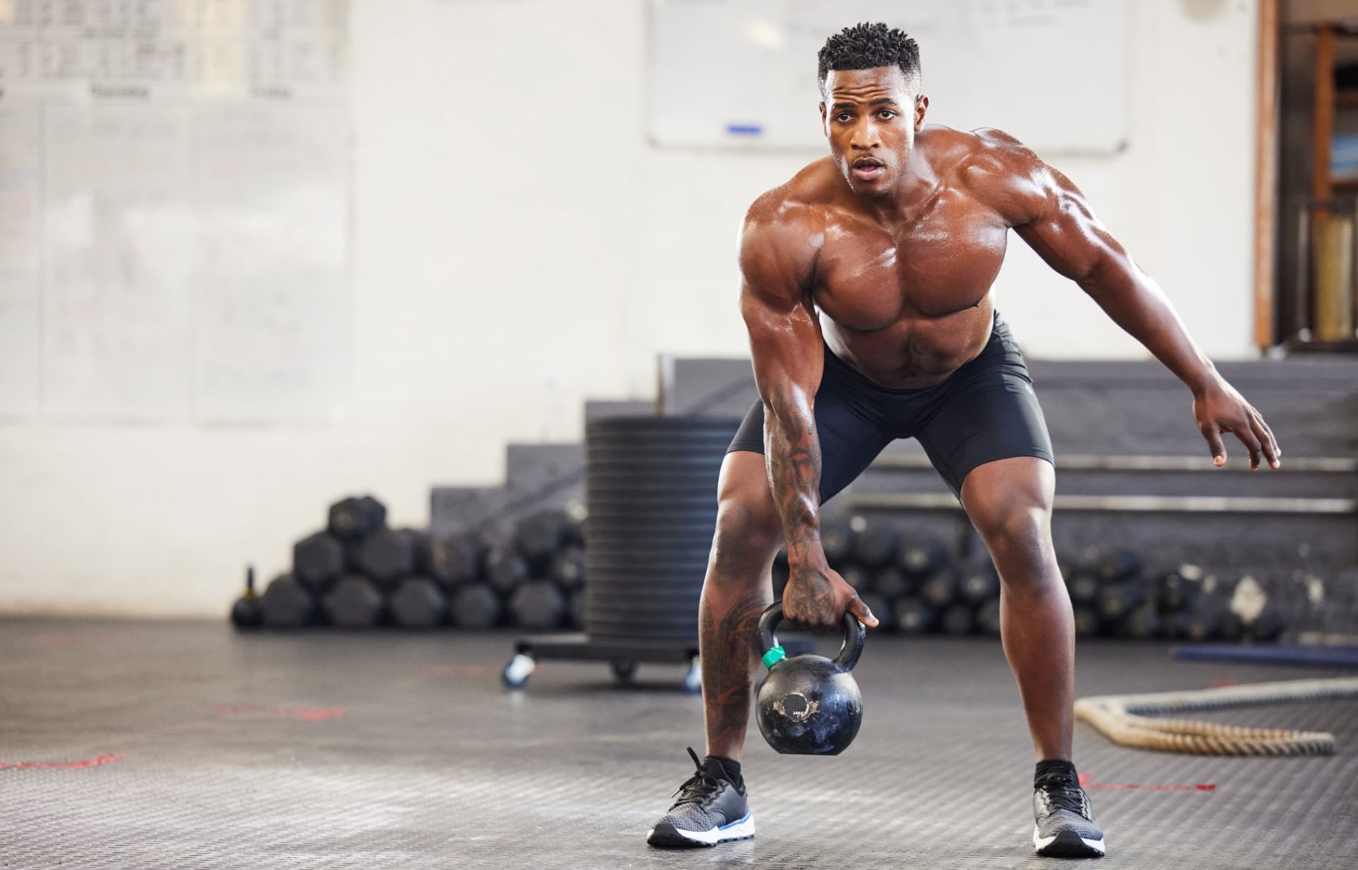 Athlete mid kettlebell swing on a gym floor with dumbbells and plates behind
